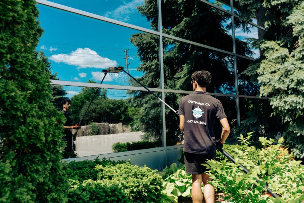 Outwash technician cleaning a glass wall with a water-fed pole beside a garden